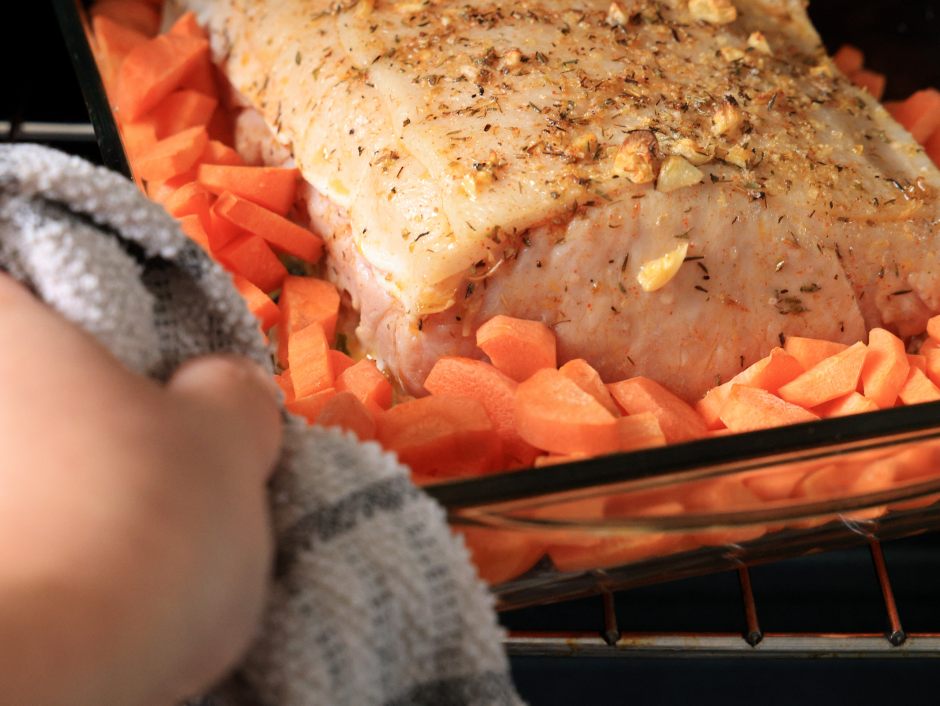 Uma pessoa usando um pano de prato retira do forno uma assadeira de vidro com um lombo suíno temperado com ervas e alho, cercado por uma grande quantidade de cenouras picadas.