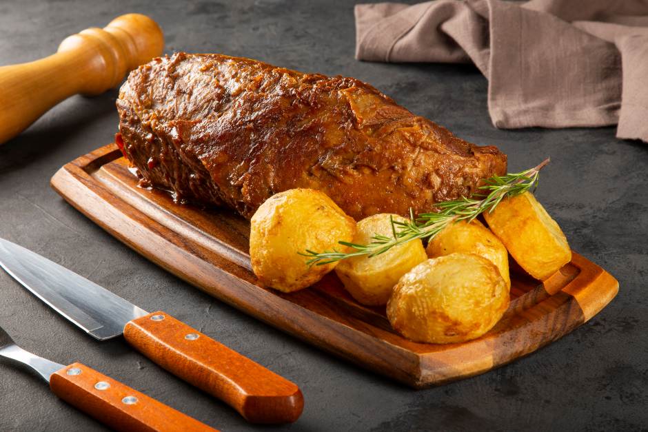 Peça inteira de lagarto assado e dourado servida em uma tábua de madeira com batatas assadas, um ramo de alecrim, faca e garfo ao lado sobre uma superfície escura.