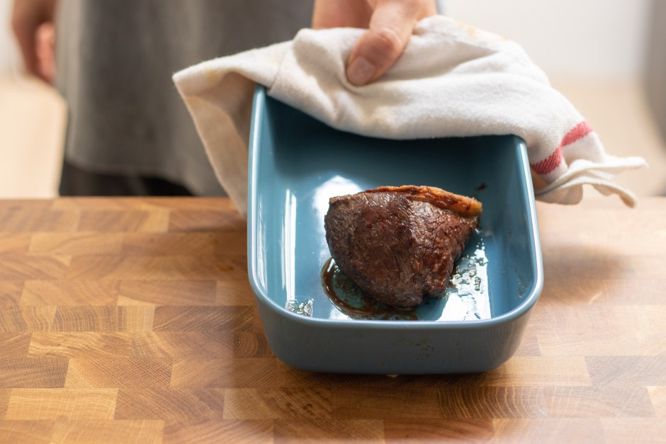 Mão de uma pessoa segurando uma travessa retangular azul com um pano de prato branco, contendo um pedaço de carne assada suculenta sobre uma bancada de madeira.