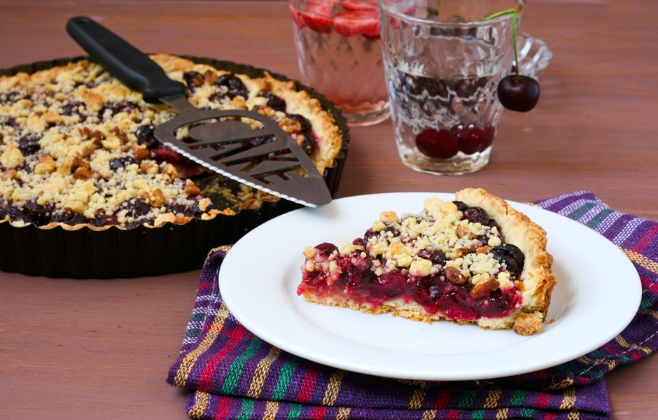 Imagem de uma torta de frutas vermelhas com cobertura de crumble, acompanhada de duas taças de bebida com frutas vermelhas e cereja, em uma mesa de madeira.