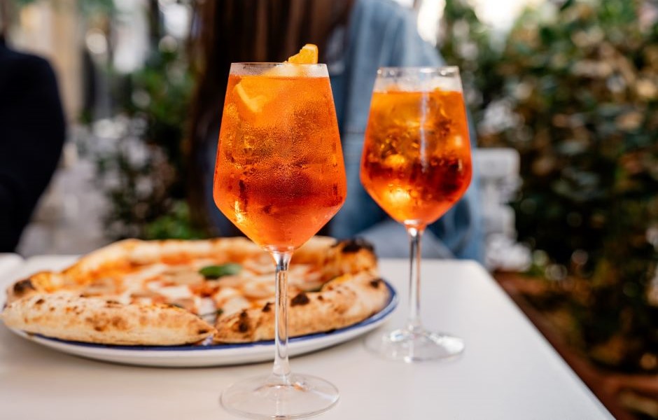 Duas taças de aperol spritz com fatias de laranja, acompanhadas de uma pizza em um ambiente ao ar livre, perfeito para momentos de lazer e convivência.