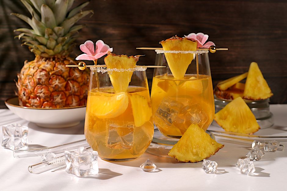 Dois copos de drink de abacaxi com gelo e decoração de flor rosa, ao fundo uma abundante fruta abacaxi e pedaços de abacaxi ao redor, criando uma atmosfera refrescante e tropical.