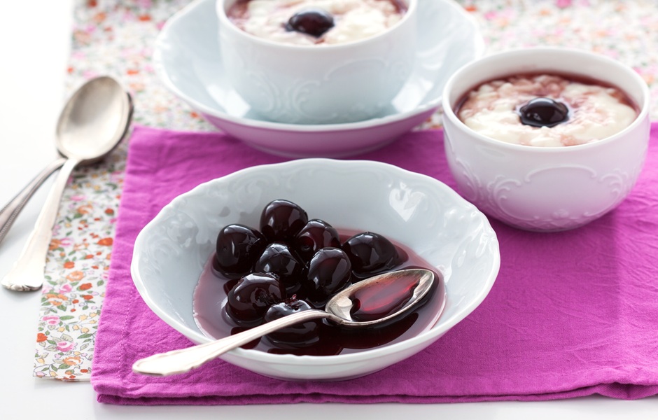 Imagem de cerejas ao vivo em uma tigela de porcelana, acompanhadas de duas taças de sobremesa de iogurte com cerejas cherry e uma colher de sobremesa ao lado, sobre uma toalha roxa com fundo floral.