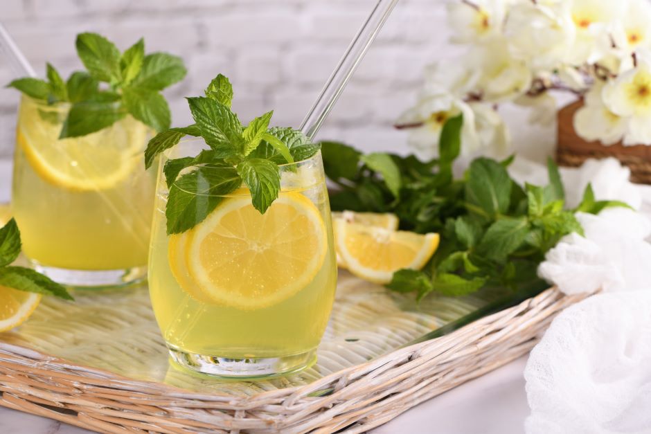 Dois copos de limonada com fatias de limão e folhas de hortelã, servidos em um ambiente decorado com flores brancas e folhas verdes, ideal para refrescar-se.