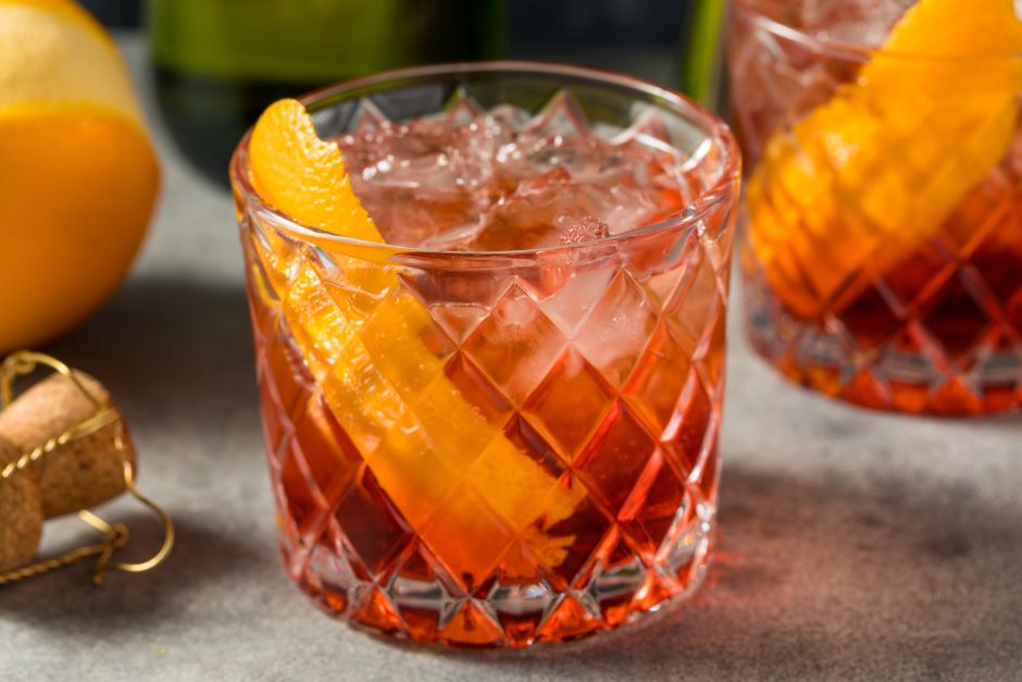 Copo de drink com gelo, fatias de laranja e uma bebida de cor rosa em um copo de vidro decorado, ao lado de outra bebida semelhante, com fundo de laranja e vegetais verdes.