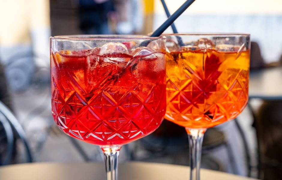 Dois copos de drinks coloridos, um vermelho e um laranja, com gelo e canudos em um ambiente ao ar livre, ideal para consumo de bebidas refrescantes.