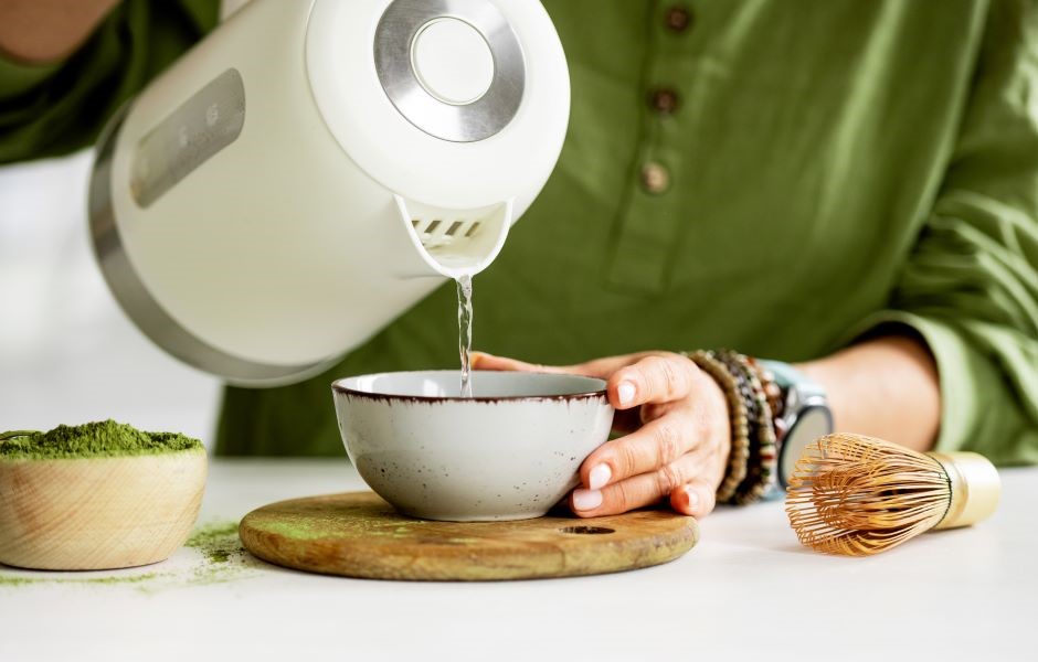 Pessoa preparando chá matcha com utensílios tradicionais, incluindo batedor de chá e tigela, em ambiente de cozinha natural e rústica.