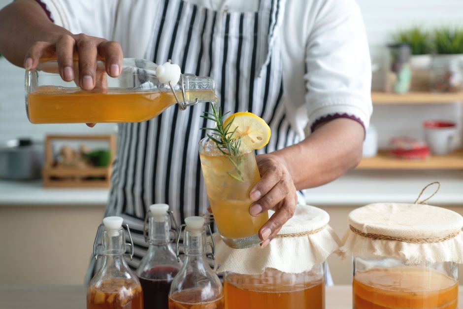 Homem preparando um drink com frutas e ervas, enchendo um copo com uma bebida refrescante de cor âmbar, em uma cozinha moderna.